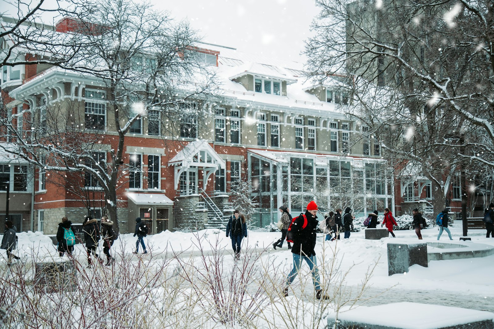 冬日雪天，穿着厚重冬装的学生们走过一座砖红色的学校建筑前，雪花纷飞